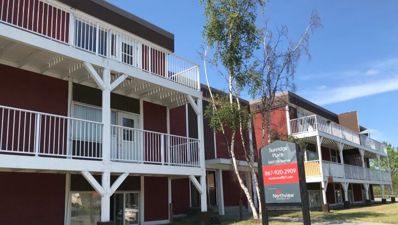 The Sunrdige Place apartment building in summer, with red paint and white balconies and a sign in front.