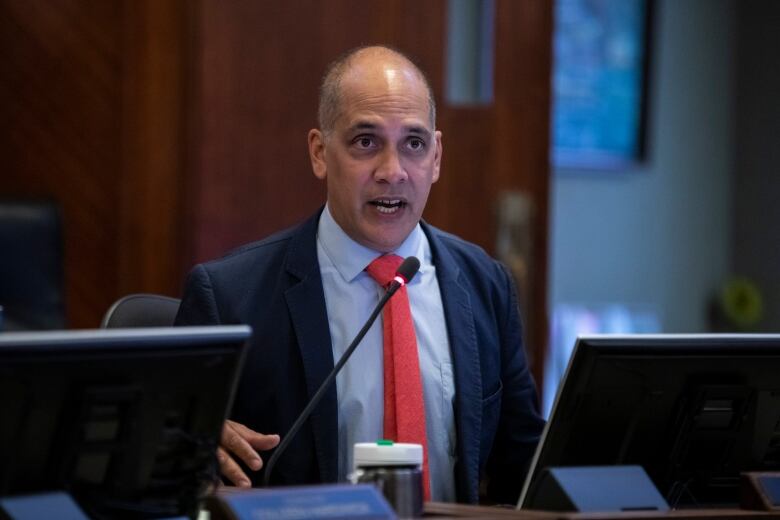 A Black man wearing a red tie speaks at a council meeting while seated.