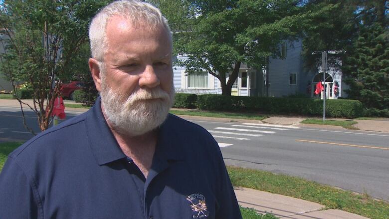 A white man with a white hair and beard wears a navy polo, standing outside near a street with a crosswalk behind him