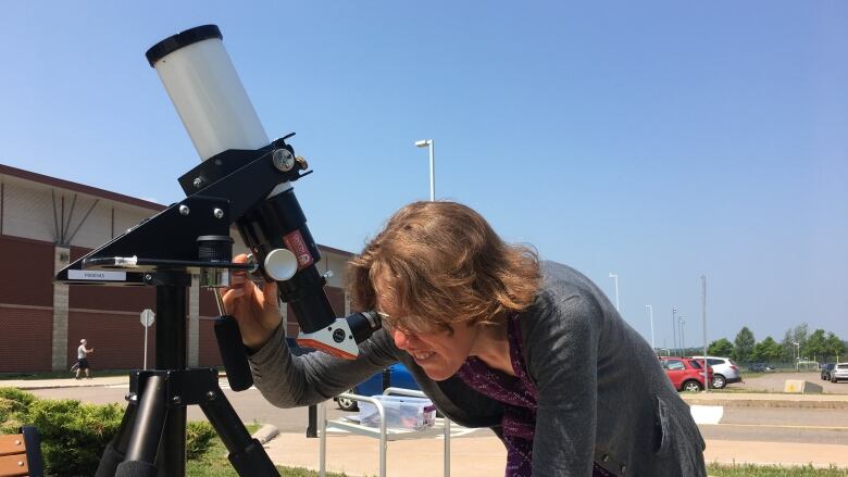 A woman with shoulder-length reddish hair and eyeglasses looks through a telescope on a university campus