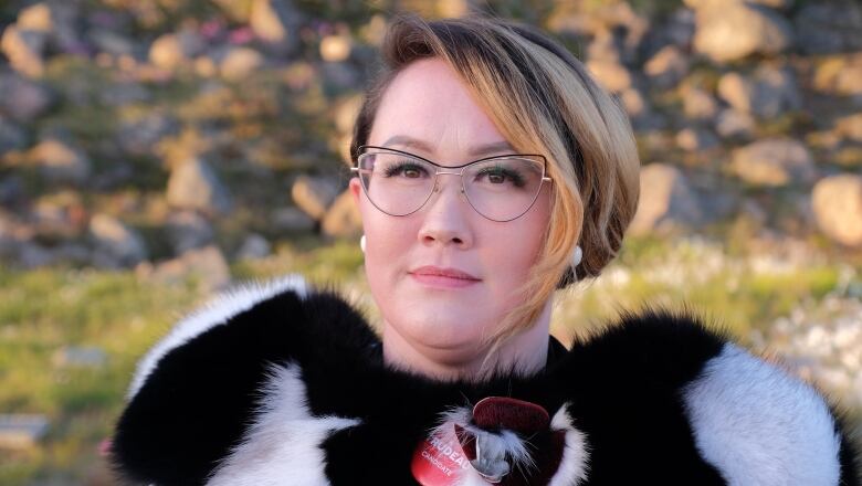 A woman with short hair and glasses wearing a fur vest poses for a photo 