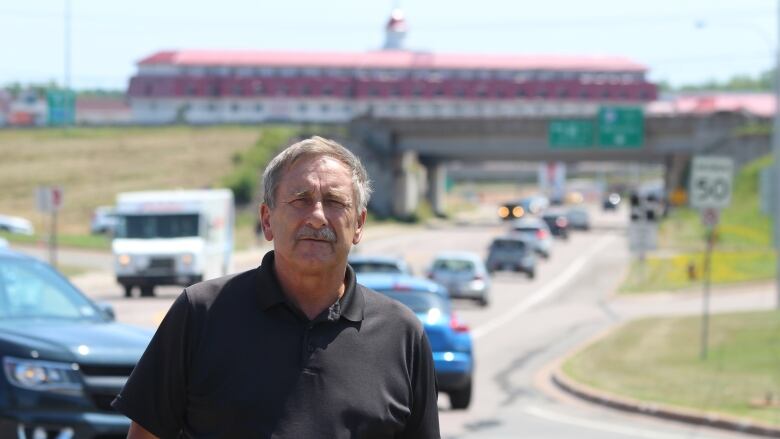 A man with short grey hair standing in front of a busy roadway with a highway overpass and a red-roofed building in the background.