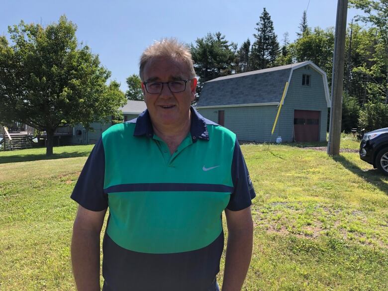 Man wearing glasses, in a green and blue golf shirt, standing in front of a house and small barn.