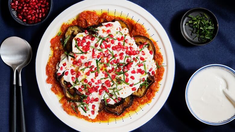 overhead shot of a white plate on a dark blue surface. the plate has eggplant borani on it, with a base of tomatoes, slices of eggplant, a layer of yogurt and topped with pomegranate arils. 