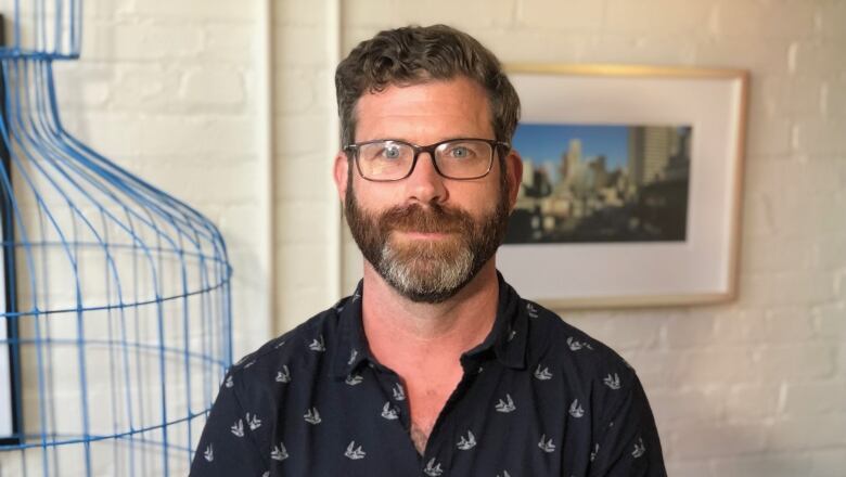 A man with dark, curly hair and a thick beard stares at the camera. He's wearing glasses and a button-up shirt. Behind him is a brick wall painted white.