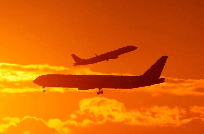 The silhouettes of two planes can be seen passing each other in an orange sky. 