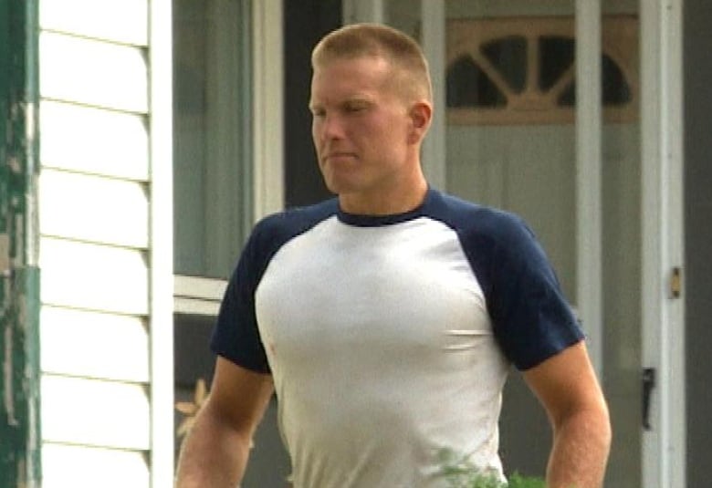 A man with blonde hair cut short in a white t-shirt carries a bunch of carrots.