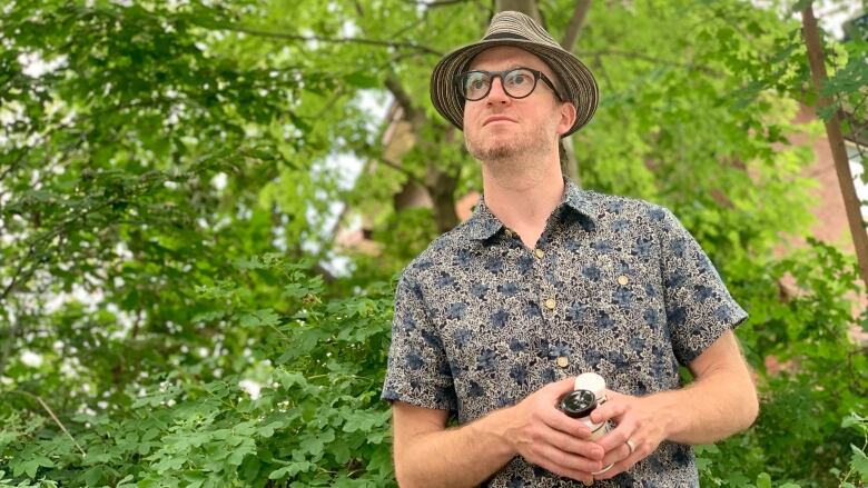 A middle aged white man stands in a park, against a backdrop of greenery, holding pill bottles.