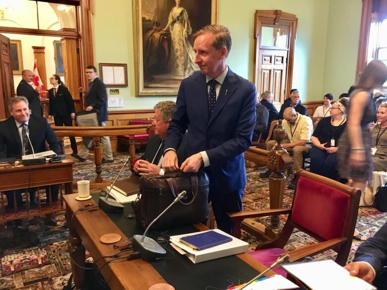 A man stands in a room with other people milling about. He has his hands on the top of a briefcase.