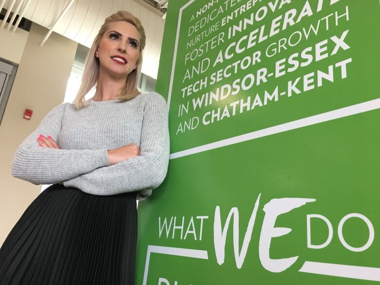 A woman with blonde hair wearing a grey shirt and black pants standing beside a green banner with white text