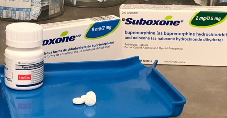 White Suboxone pills are displayed on a blue tray with a bottle and small boxes of the drug in the background.