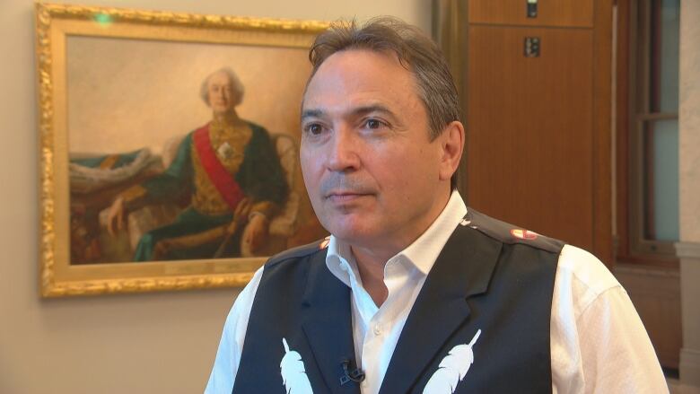 A man wears a white shirt and a dark vest with images of feathers.