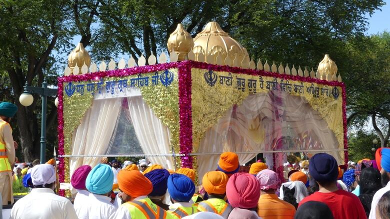 A float with a golden domed peak and white curtains passes through a crowd. Several people in the crowd are wearing orange and blue turbans. 