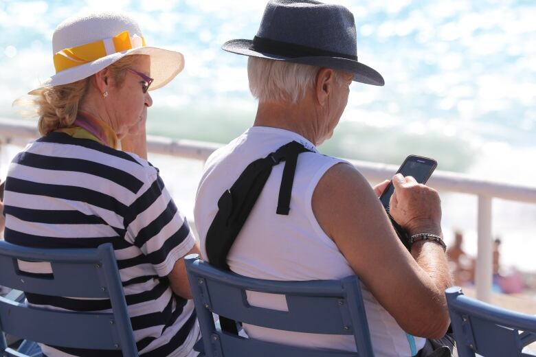 An elderly man scrolls on a mobile phone while sitting on a bench near a beachfront. He sits next to an elderly woman. 