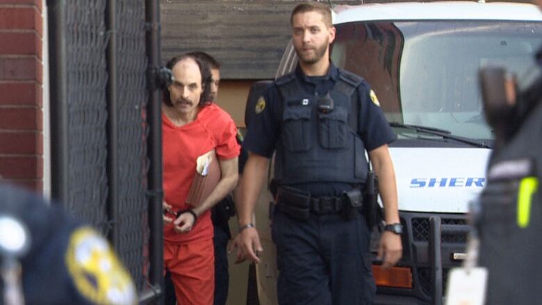 A man in an orange jumpsuit is led by uniformed peace sheriffs.