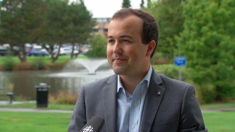 A man in a sport coat stands for a news interview outside with a park in the background.