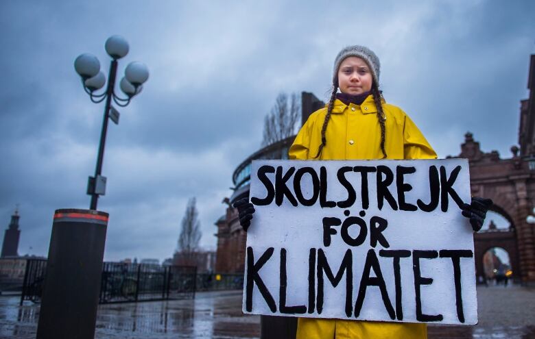 Greta Thunberg wears a grey hat and a yellow rain jacket. She's holding a sign that reads 