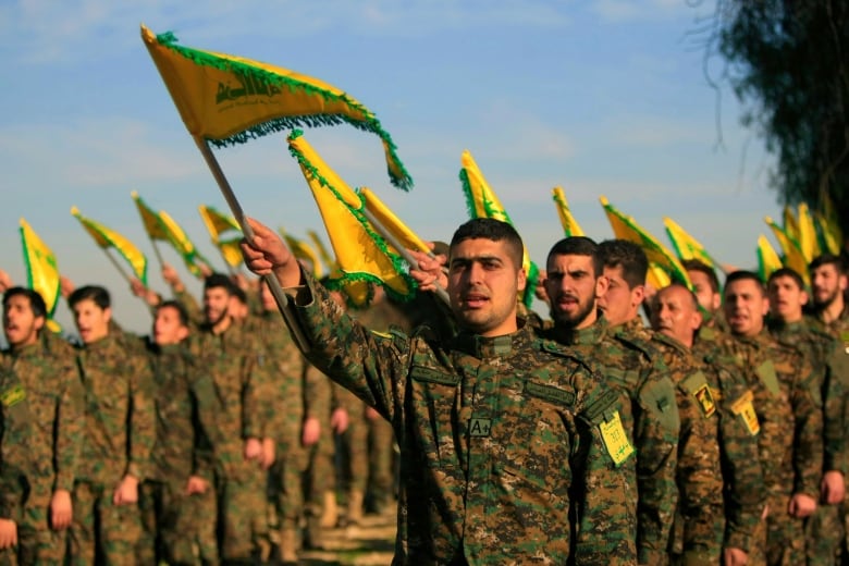 Dozens of men in military uniforms, waving green-and-yellow flags, stand in formation.