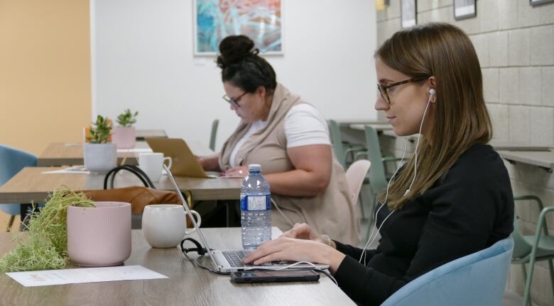 Two women sit at tables, working on laptops.