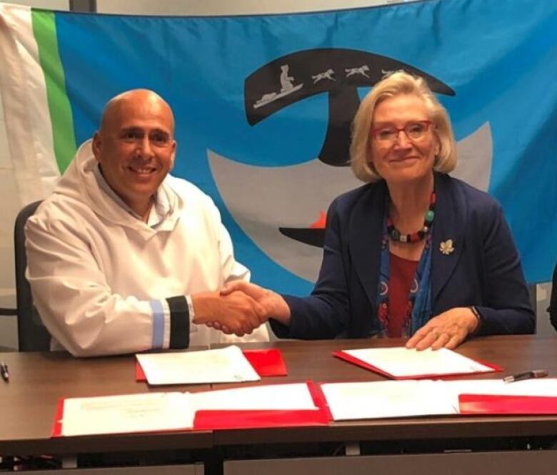 A man in a white top shakes hands with a woman in a blue jacket. They are sitting at a table with papers on it.