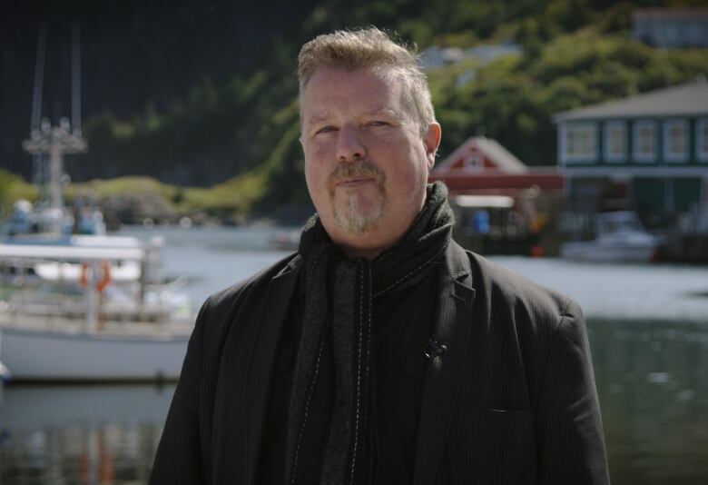 A man with a goatee stands in front of fishing buildings by a harbour.