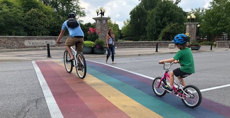 An adult and child cycle over a rainbow crossing into a park.