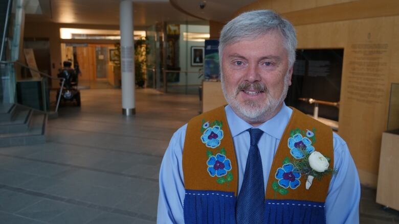 A white man with grey hair smiles at the camera. He is wearing a beaded vest. 