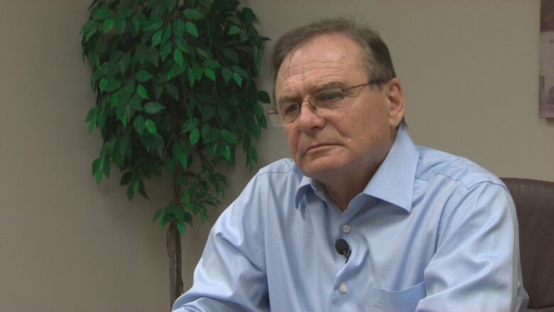 A older white man with brown hair is wearing rectangular glasses and a blue button up shirt.