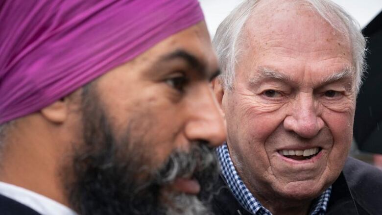 A man with white hair looks on as a man in a purple Turban greets people in a market square.