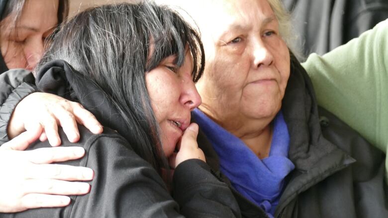 A woman wipes her face as she leans against an older woman who has her arm around the younger woman.