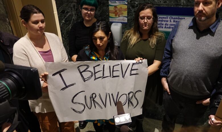 A group of people stands with a sign reading 'I believe survivors.'