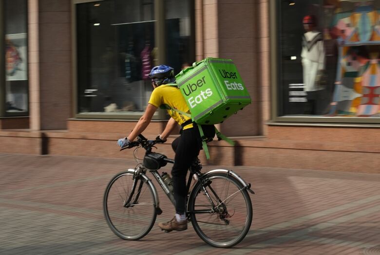 A man rides a bicycle on a sidewalk, passing several storefronts. On his back is a large green box that says 'Uber Eats.'