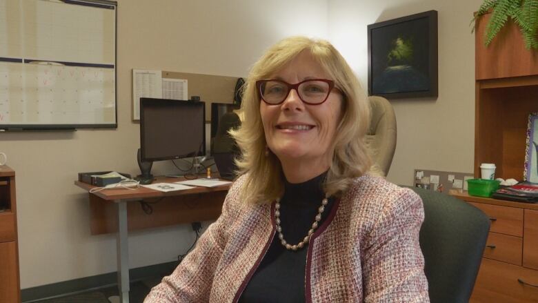 Woman in glasses and a pink tweed blazer sits at a desk and smiles into the camera.