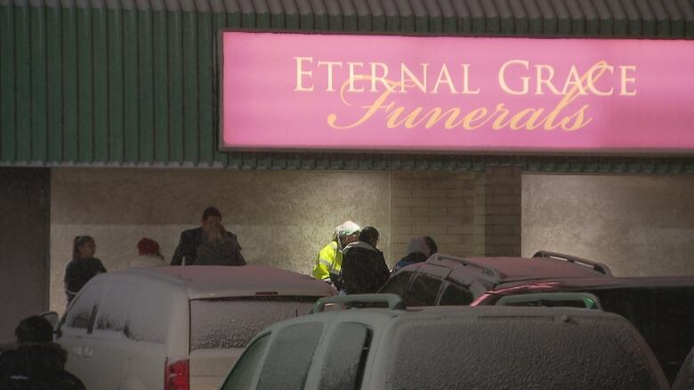 The tops of snow-covered vehicles are seen in a parking lot, with people standing beyond them near a building with a sign that says 