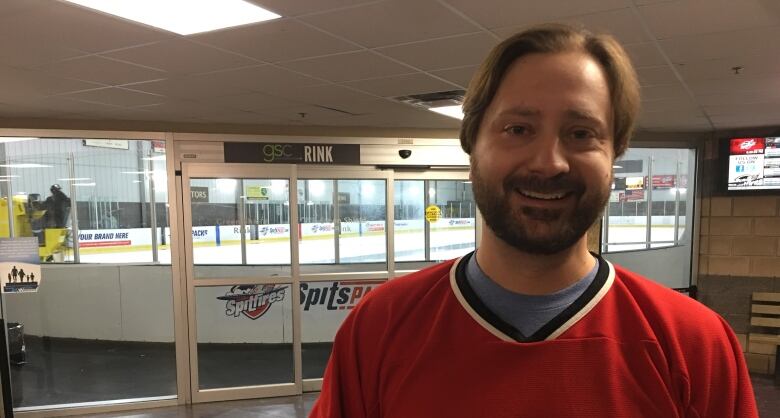 A man with brown hair and a beard wearing a hockey jersey in front of a hockey arena