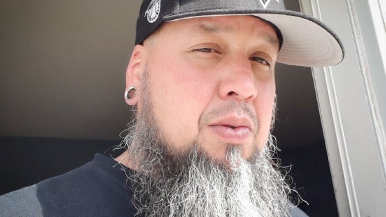 A man wearing a black ballcap and silver earring with a long salt-and-pepper beard and thin moustache looks at the camera.