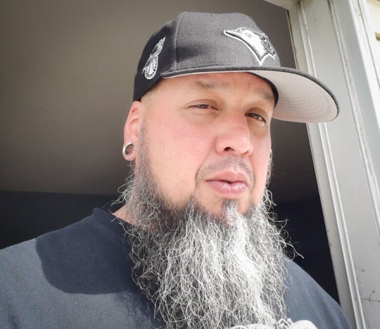 A man wearing a black ballcap and silver earring with a long salt-and-pepper beard and thin moustache looks at the camera.