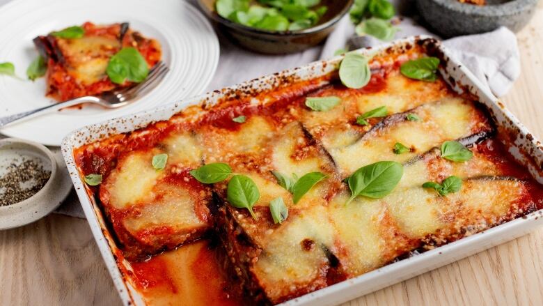 overhead shot of a white baking dish with eggplant parmesan in it. it's on a wooden table. a slice is missing from the dish and is sitting on a white plate behind the baking dish. 