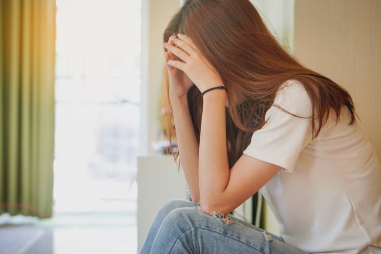 A female teen sits in a bright room, hiding her face in her hands and behind her long hair.