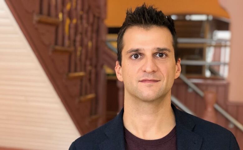 A clean-shaven man with dark hair stands in front of a brown staircase.