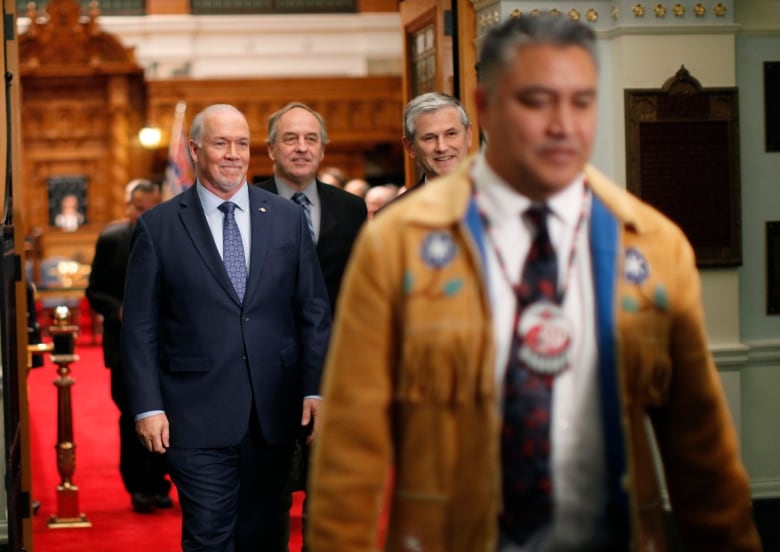 People in suits walk down a hall behind an Indigenous man in a buckskin jacket.