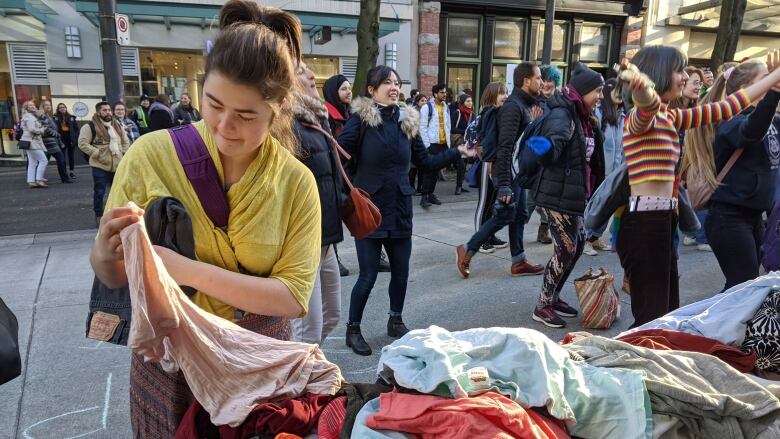 A woman picks up an item of clothing from a table on a sidewalk as many other people gather in the background.