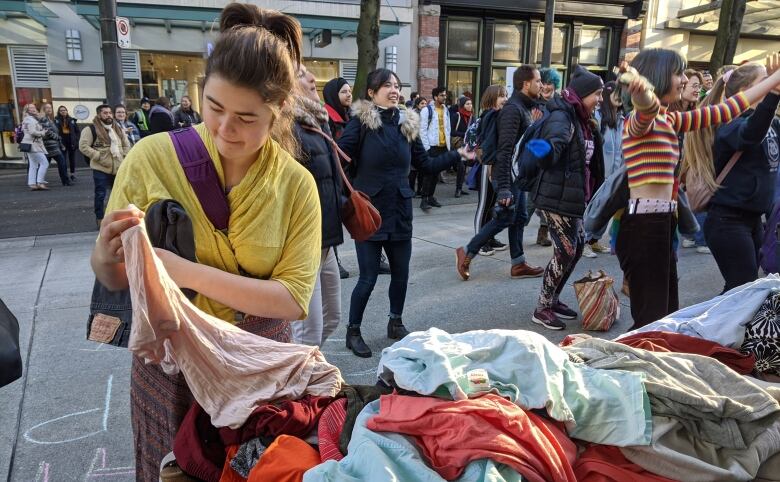 A woman picks up an item of clothing from a table on a sidewalk as many other people gather in the background.