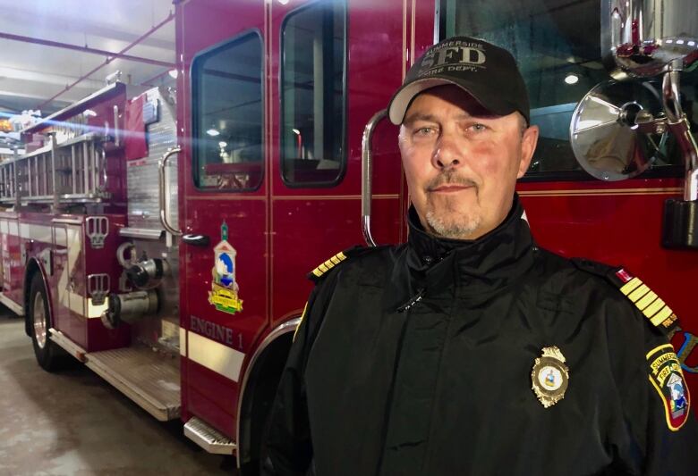 A bearded man in a black fire department uniform stands in front of a fire truck. 