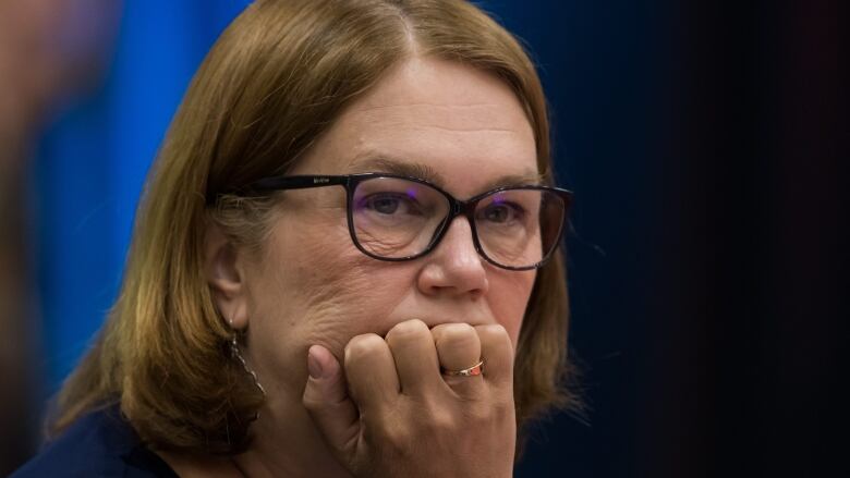 A woman with light brown hair and black glasses places her hand on her chin while looking seriously onwards.