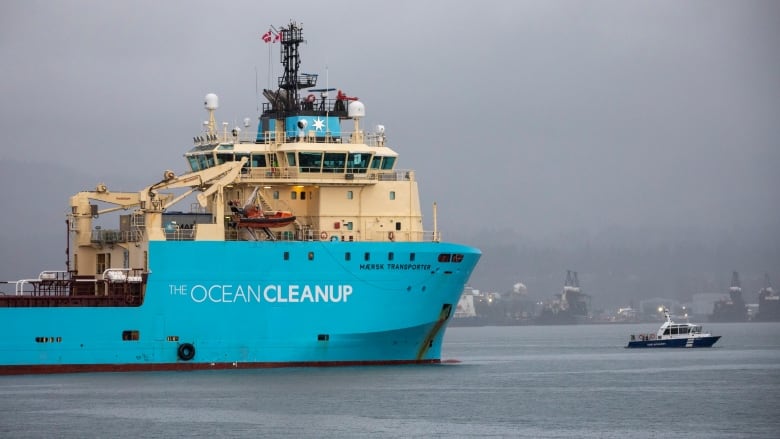 The front of a large blue freighter with the words Ocean Cleanup plies the waters of a harbour with other ships in the background on a foggy day.