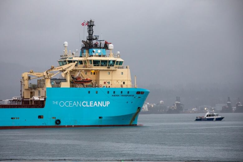 The front of a large blue freighter with the words Ocean Cleanup plies the waters of a harbour with other ships in the background on a foggy day.