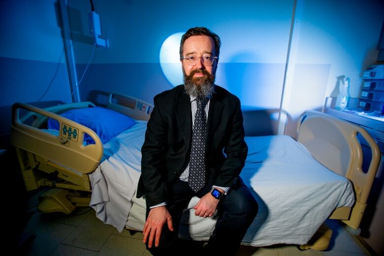 A white man with a medium-length salt-and-pepper beard sits on a hospital bed. He is wearing a dark suit with a patterned tie.