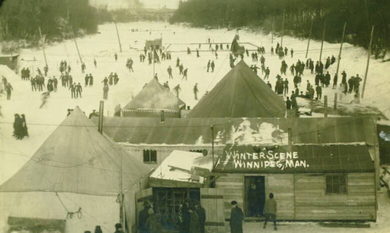 Black and white photo of people skating on a river. A wooden shack and attached tent is in the foreground.