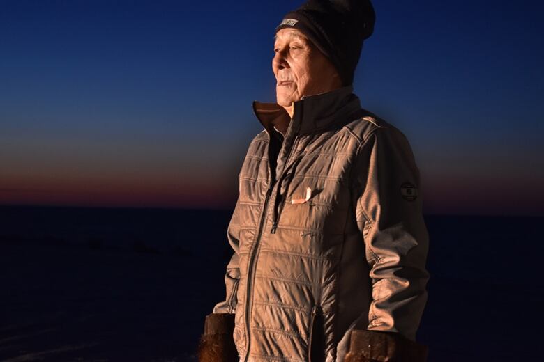 A man with a brown coat, brown mitts, and a black hat stares into light against a dark outdoor background.